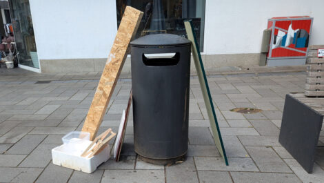 Ein öffentlicher Mülleimer in einer Fußgängerzone, an den Regalteile und eine Werbetafel aus einem Ladengschäft angelehnt sind.