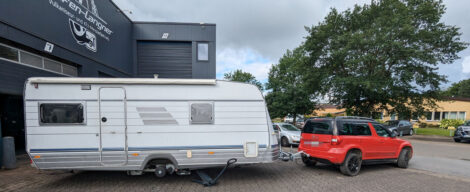 Ein weißer Wohnwagen hinter einem roten Auto. Das recht Rad des Wohnwagen ist demontiert.