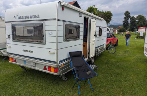 Ein Wohnwagen mit offener Tür und einen Campingstuhl davor auf einer Rasenfläche.