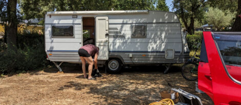 Ein Wohnwagen auf einem Campingplatz mit trockenem Boden. Davor steht gebückt ein Mann und hantiert mit irgendwas herum