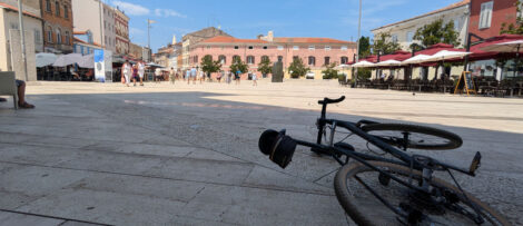 Ein Fahrrad liegt flach im Schatten eines Gebäudes auf dem Boden eines großen Platzes.