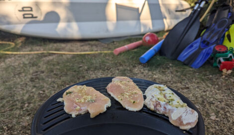 Drei Stückchen Hähnchenbrust auf einem Gasgrill. In Hintergrund ist unscharf ein Paddelbrett zu erkennen.