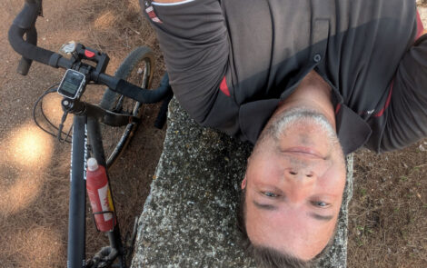 Ein Mann liegt "auf dem Kopf" auf einer Mauer und schaut in die Kamera. Links neben der Mauer sieht man ein angelehntes Fahrrad.