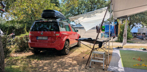 Zwischen der Markise eines Wohnwagen und der Dachreeling  eines Autos ist eine Plane gespannt und spendet so Schatten. 