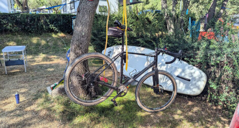 Ein Fahrrad ist am Sattel mit einem Spanngurt an einem Baum aufgehängt. Das Vorderrad berührt den Boden und das Hinterrad schwebt frei in der Luft.