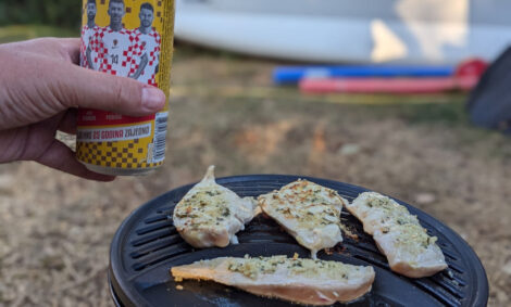 In einer Hand wird eine Dose kroatisches Bier schräg über einen Minigrill mit 4 Hähnchenschnitzeln gehalten. Im Hintergrund ist unscharf ein PAddelbrett zu sehen.