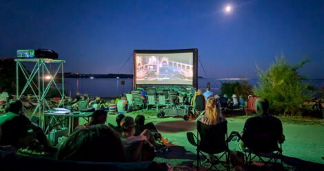 Eine aufblasbare Kinoleinwand vor dem Meer und einem mitternachtsblauem Himmel. Davor sitzen viele Zuschauer in Campingstühlen mit dem Rücken zur Kamera und verfolgen den Film.