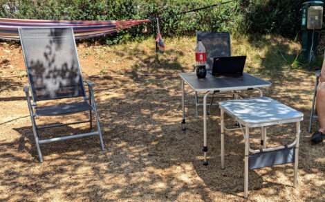 Ein Campingstuhl neben zwei Campingtischen auf trockenem, braunem Boden. Im Hintergrund ist eine grüne Hecke zu sehen.