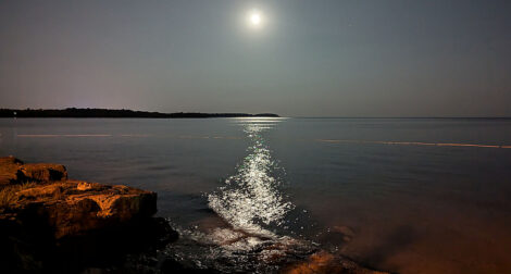 Der Mond spiegelt sich auf einer glatten Meeresoberfläche. Am Horizont sieht man flach eine Landzunge.