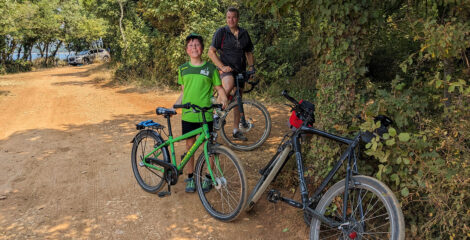 Ein Junge und ein Mann stehen neben ihren Fahrrädern und schauen in die Kamera. Davor lehnt ein Fahrrad an einem Baum.