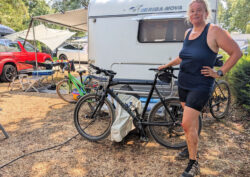 Eine Frau steht neben einem Fahrrad, welches an einem Wohnwagen lehnt und hat die Hand auf dem Sattel abgestützt. Das linke Bein ist verschrammt