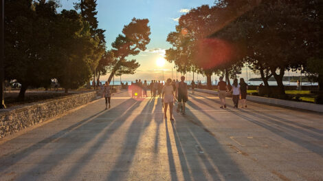 Menschen auf einer Promenade mit Bäumen rechts und links vor einem orangenen Sonnenuntergang.