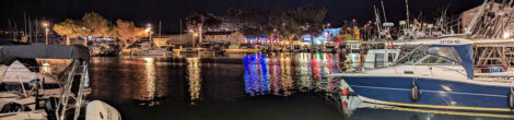 Ein Hafen mit einigen Booten bei Nacht. Auf der gegenüberliegenden Seite strahlen bunte Lichter, die sich im Hafenbecken spiegeln.