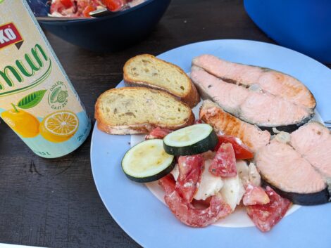 Ein Teller mit zwei gegrillten Scheiben Lachs, etwas Tomatensalat mit Gurke, zwei gerösteten Weißbrotscheiben und einer Dose Radler.