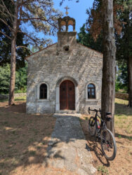 Eine kleine Kapelle aus Natursteinen unter Bäumen. Im Vordergrund ist ein Fahrrad an einen Baum gelehnt.