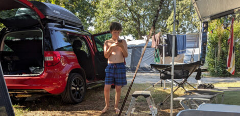 Ein Auto mit einer offenen Heckklappe auf einem Campingplatz. Daneben steht ein kleiner Junge und schaut auf sein Handy.