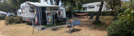 Ein Wohnwagen auf einem Campingplatz mit verdorrtem Gras und einigen kleinen Eichenbäumen. Im Vordergrund steht ein Campingstuhl, auf dem eine Solarzelle liegt.