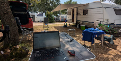 Stellplatz auf einem Campingplatz mit einem Wohnwagen mit ausgefahrener Markise, einem PKW mit offener Heckklappe und einem Campingtisch im Vordergrund, auf dem ein aufgeklappter Laptop steht.