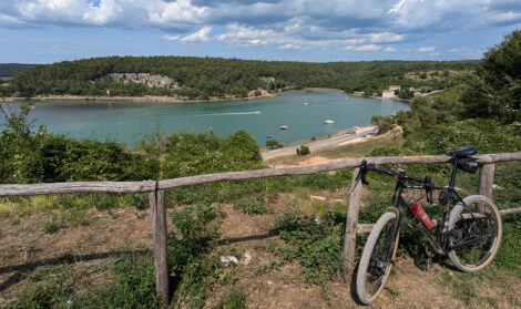 Ein Gravelbike lehnt an einem Holzzaun aus runden Naturstämmen oberhalb einer Bucht, auf deren Wasser einige Sportboote fahren. Im Hintergrund ist ein bewaldeter Hügel unter einem blauen Himmel mit einigen weißen Wolken zu sehen.