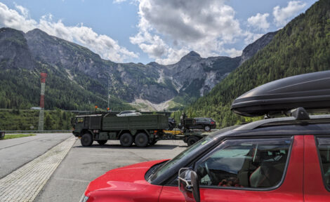 Ein Gebirgsmassiv. Im Vordergrund ein roter PKW und etwas dahinter ein Militärtransporter