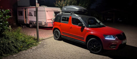 Ein Wohnwagen mit offener Tür vor einen Haus. Im Vordergrund steht das Zugfahrzeug und wird von einer Straßenlaterne beleuchtet.