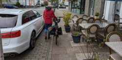 Ein Radfahrerin schiebt zwischen einem parkenden Auto und der fast direkt angrenzenden Außenbestuhlung einer Gastronomie auf einem viel zu schmalen Stück Gehweg.