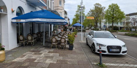 Ein Gehweg mit opulenter Außenbestuhlung einer Gastronomie und einer direkt daran angrenzenden Parkfläche für KFZ. Dazwischen ist ein sehr schmaler Streifen frei, den zu Fuß Gehende nutzen können.