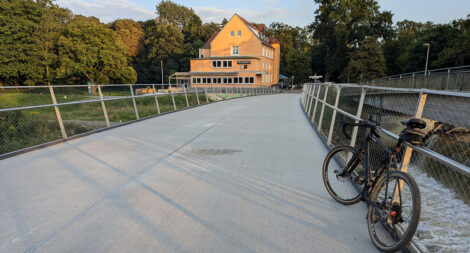 Am Geländer einer Brücke für Fahrradfahrende und zu Fuß Gehende lehnt ein Fahrrad am Geländer. Am Ende der Brücke ist ein gelbes Haus zu sehen.