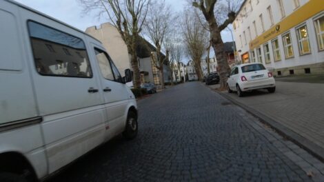 Auf einer Kopfsteinpflasterstraße überholt ein weißer Van einen Radfahrer mit zu wenig Seitenabstand.