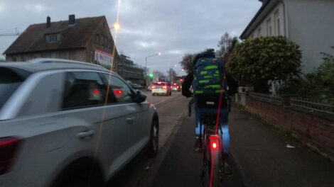 Ein sehr schmaler benutzungpflichtiger Hochbordradweg mit links angelegten Parkbuchten. Man sieht einen jungen Schüler auf seinem Fahrrad, welches die Radwegbreite komplett ausnutzt.