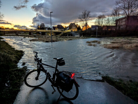 Ein überfluteter Radweg durch eine Mulde. Im Hintergrund ist eine beleuchtete Kfz-Brücke zu sehen und am Himmel ziehen schwere, schwarze Wolken entlang.