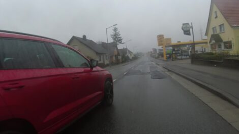 Ein rotes Auto wird mit zu wenig Abstand an einem fahrradfahrer vorbei gesteuert. Die Straße ist feucht und abschüssig.