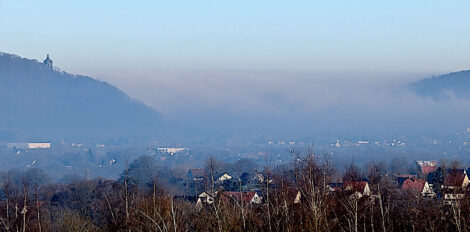 Zwischen zwei Hügelketten quillt Nebel durch das Tal. Im Vordergrund sieht man Bäume und Häuser.