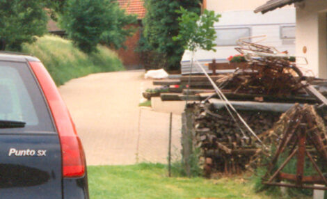 Links sieht man einen kleinen Teil der rechten Seite eines Autos im Hintergrund ist ein Wohnwagen zu sehen und rechts ein Schuppen. In der Mitte dieses Bildes ist ein junger Baum neben einem Holzstapel zu sehen.