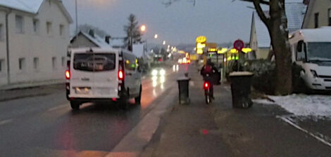 Ein Junge fährt morgens im Halbdunklen mit seinem Fahrrad auf einem kombinierten Geh- und Radweg zwischen zwei Mülltonnen hindurch.