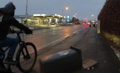 Ein Junge schlängelt sich  morgens im Halbdunklen mit seinem Fahrrad auf einem kombinierten Geh- und Radweg neben einer quer auf dem Radweg liegenden Mülltonne hindurch.