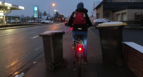 Ein Junge fährt morgens im Halbdunklen mit seinem Fahrrad auf einem kombinierten Geh- und Radweg zwischen zwei Mülltonnen hindurch.