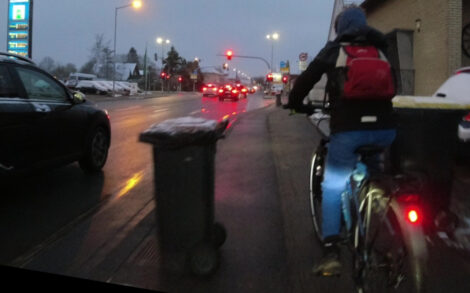 Ein Junge fährt morgens im Halbdunklen mit seinem Fahrrad auf einem kombinierten Geh- und Radweg neben einer mitten auf dem Radwegteil stehenden Mülltonne entlang