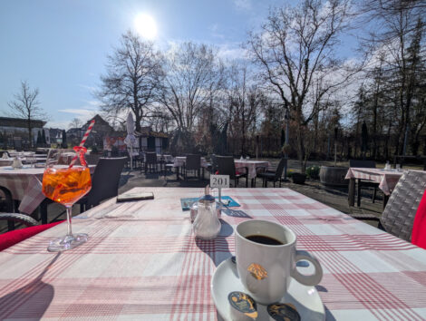 Ein Aperol-Spritz und eine Tasse Kaffee stehen auf einem sonnenbeschienenen Tisch. Im Hintergrund steht die Frühlingssonne über noch unbelaubten Bäumen.