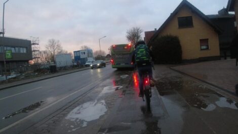 Ein Junge fährt auf seinem Fahrrad auf einem radweg, der von einem durch Pfützen fahrenden Bus komplett überflutet wurde.